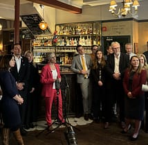 Daisy is standing in a pub, in front of the bar, delivering a speech. There are a number of people stood on either side of her.