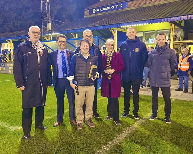 A group of people, including Daisy, are standing on a football pitch. They are all smiling and looking towards a camera, and St Albans City FC can be read on a sign on a building behind them.