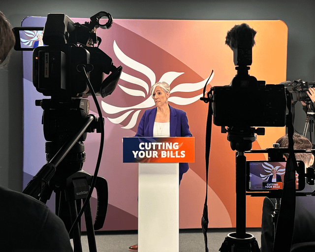 Daisy Cooper is standing at a podium, delivering a speech. The text on the podium reads 'cutting your bills'. At the forefront of the photo are some cameras, facing Daisy.