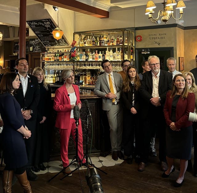 Daisy is standing in a pub, in front of the bar, delivering a speech. There are a number of people stood on either side of her.