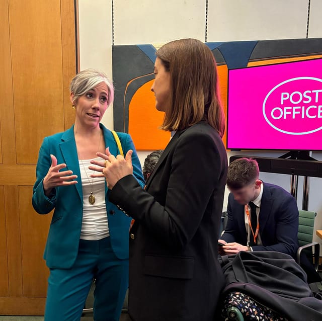Daisy Cooper MP speaking with a representative from Post Office HQ in Parliament. They are standing in a meeting room, in front of a screen showing the Post Office logo.