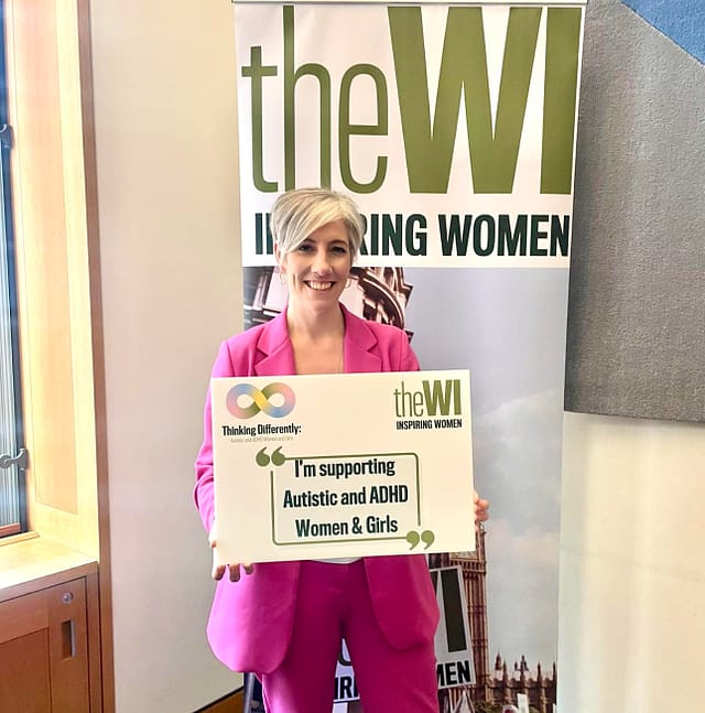 Photo of Daisy Cooper in a pink suit, holding a sign that says 'I'm supporting Autistic and ADHD Women and Girls''